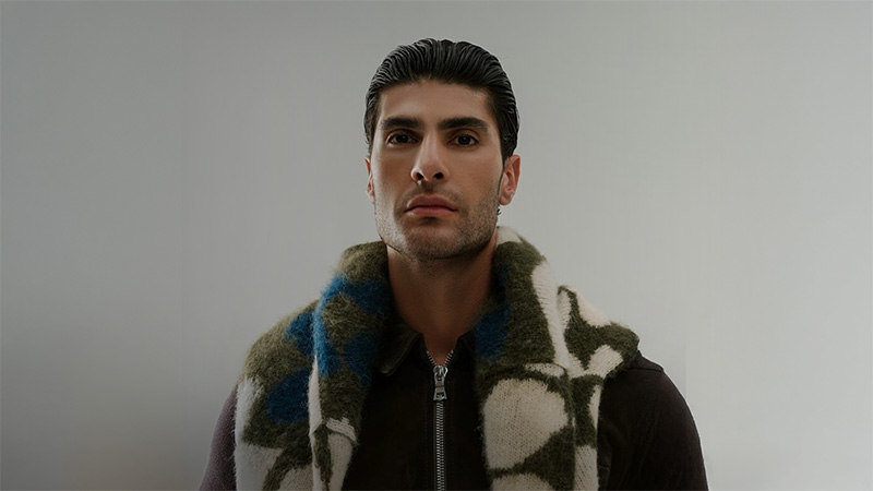 Young man with slicked-back hair wearing a dark zip-up jacket and a patterned scarf against a neutral background, projecting a confident expression.