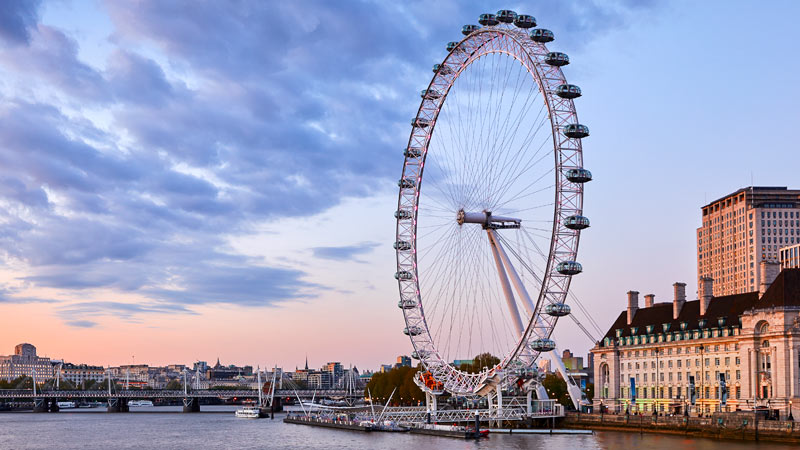 Sunset view of the London Eye by the River Thames, with a partly cloudy sky. The Ferris wheel is lit softly, reflecting tranquility and iconic cityscape.