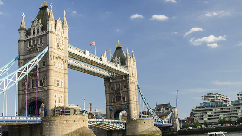Tower Bridge in London under a clear blue sky. The iconic bridge, with its two large towers and blue suspension cables, spans the River Thames.