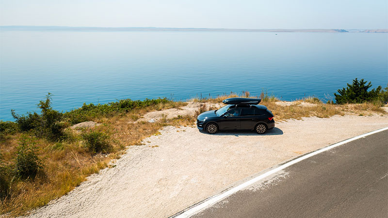 A black car with a roof box is parked on a gravel area by the sea, next to a winding road and dry grass, with calm blue water in the background.